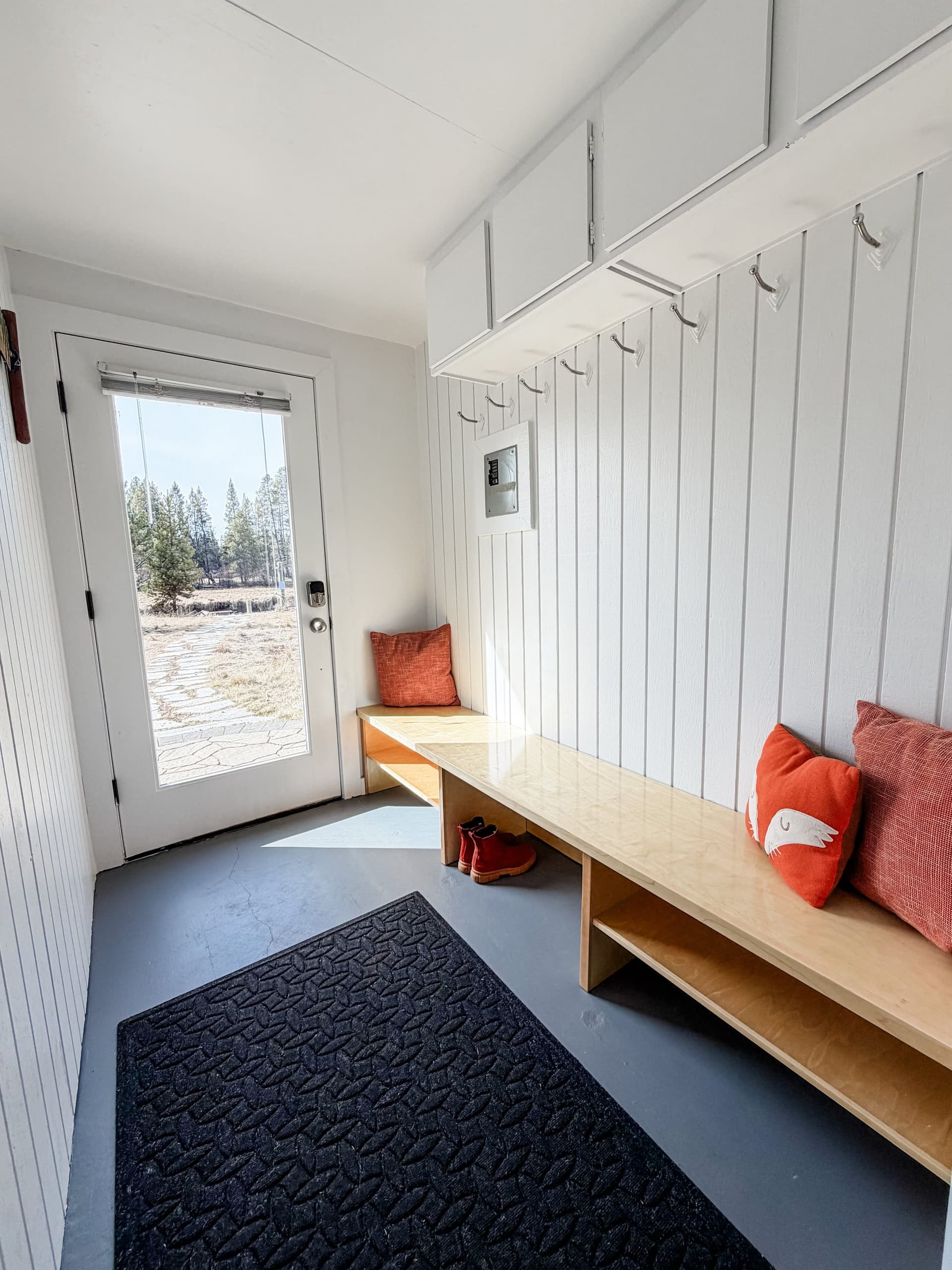 Small entryway with white paneled walls, a glass door leading outside, a wooden bench with orange pillows and red shoes.