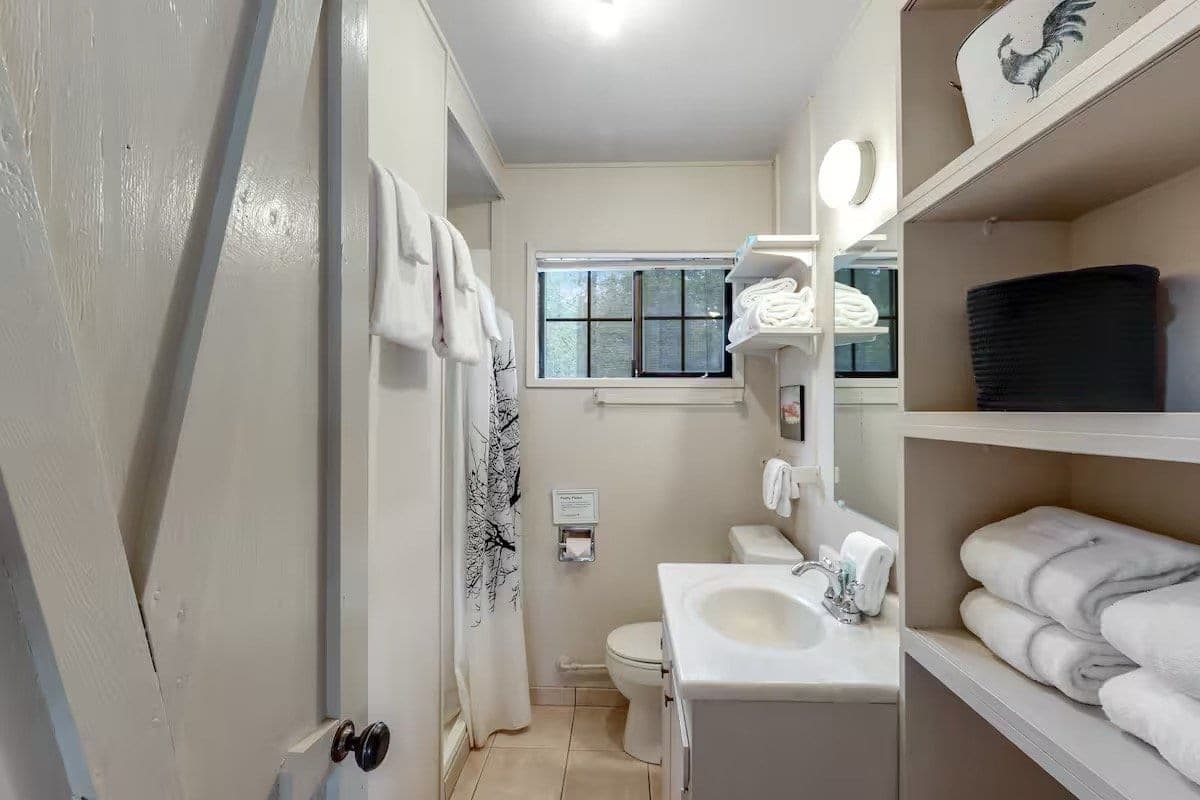 Small bathroom with white walls and beige tile floor, featuring a window with blinds, a white toilet, a white sink.