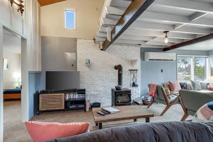 Living room with white brick fireplace, wood stove, and large windows with view of trees, furnished with colorful chairs.