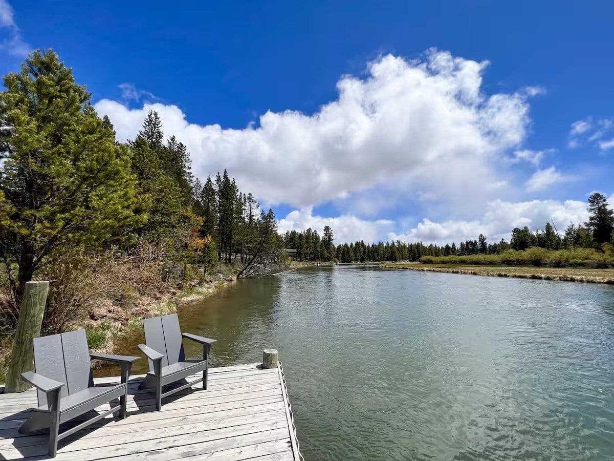 Two chairs on a wooden dock by a river with trees and a cloudy sky in the background.