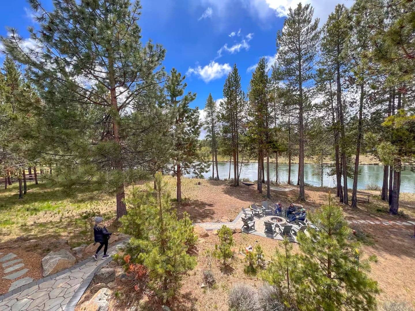 A park with tall pine trees near a river, with a stone-paved walking path, a fire pit surrounded by chairs, and a person, Deschutes Riverfront Retreat near Sunriver, Oregon