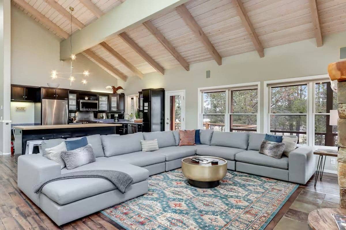 Great room with a large grey sectional on a patterned rug, an open kitchen with dark cabinets and a wall of windows, Deschutes Riverfront Retreat near Sunriver, Oregon