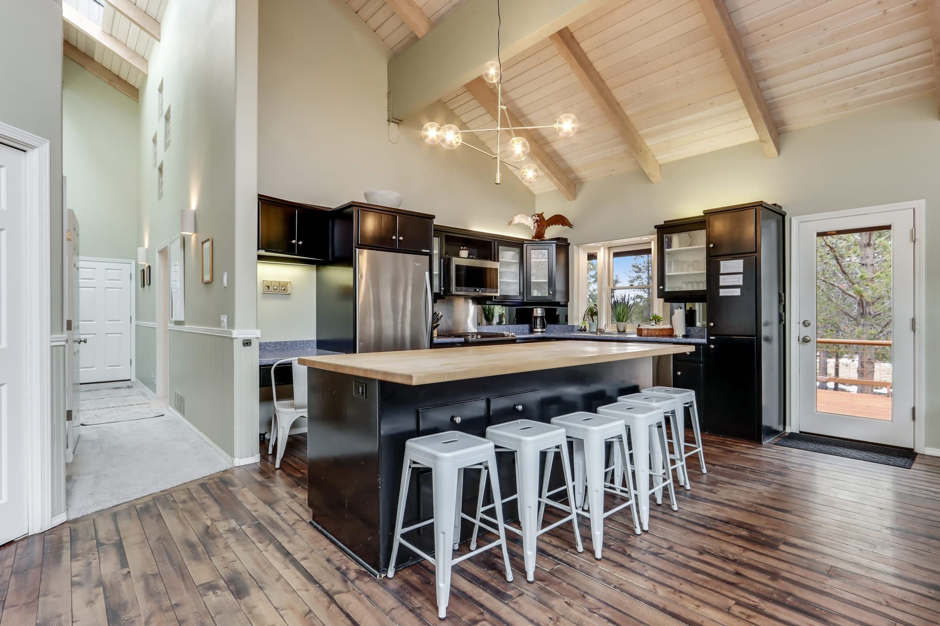 Open kitchen with a large island, five white stools, dark cabinets, stainless steel appliances, and a vaulted ceiling.