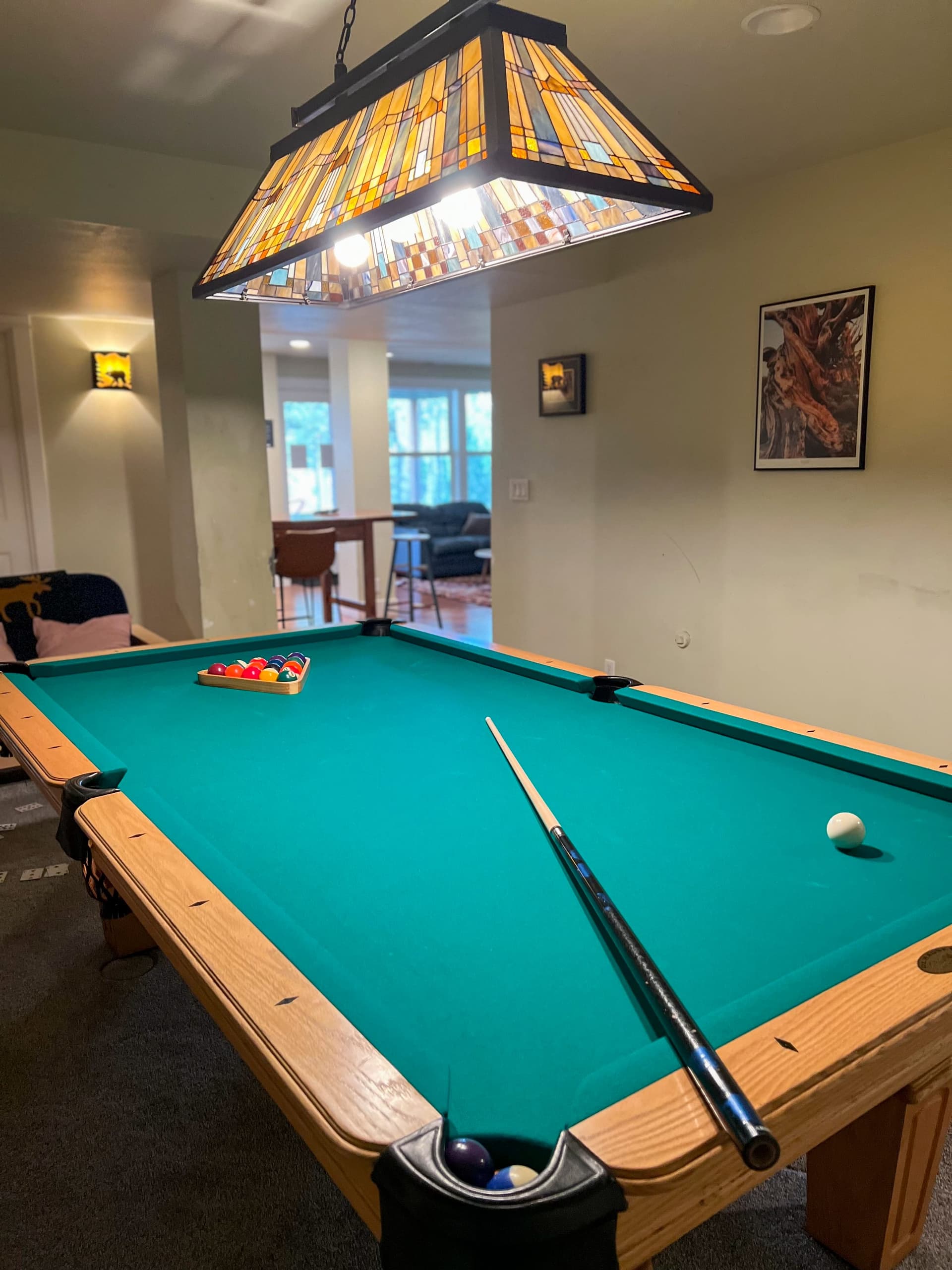 A pool table with a green felt surface, positioned under a stained glass ceiling light fixture, in a cozy living room.