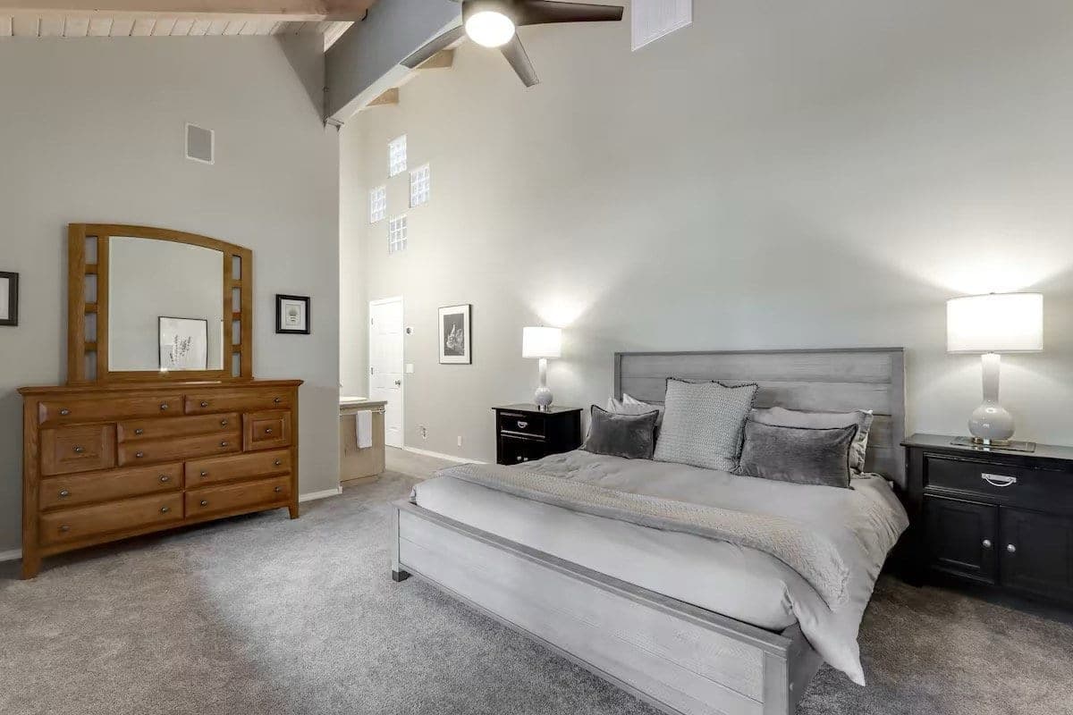 A bedroom with a large bed with gray headboard, pillows, and blankets, two nightstands with lamps, a wooden dresser.