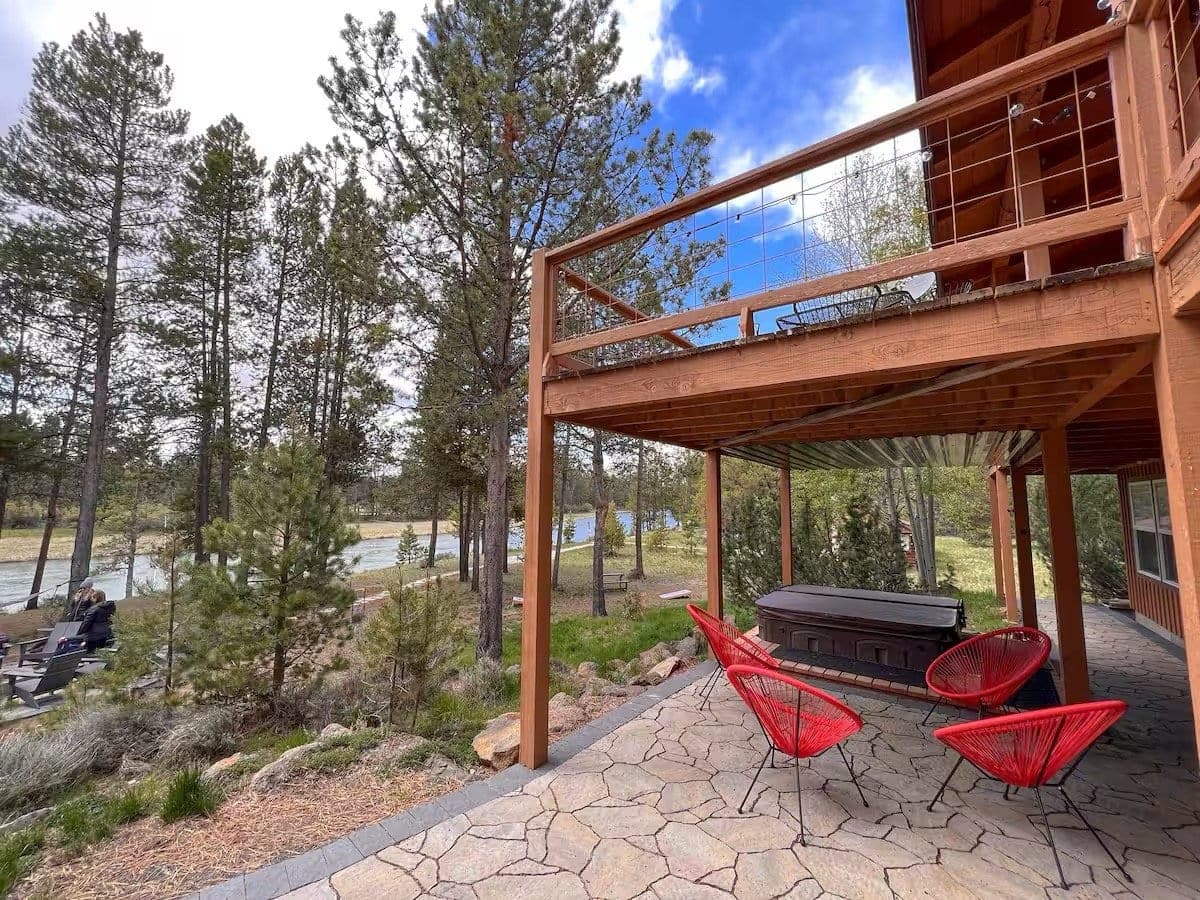 View of a wooden deck with red chairs and a hot tub, overlooking a pine forest and a river, under a partly cloudy sky.