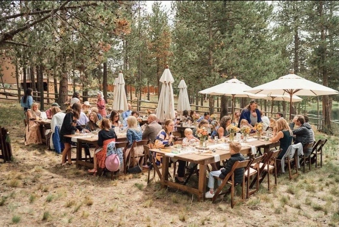 Wedding guests seated at long tables under the pines, parasols shading the far end, Deschutes Riverfront Retreat near Sunriver, Oregon