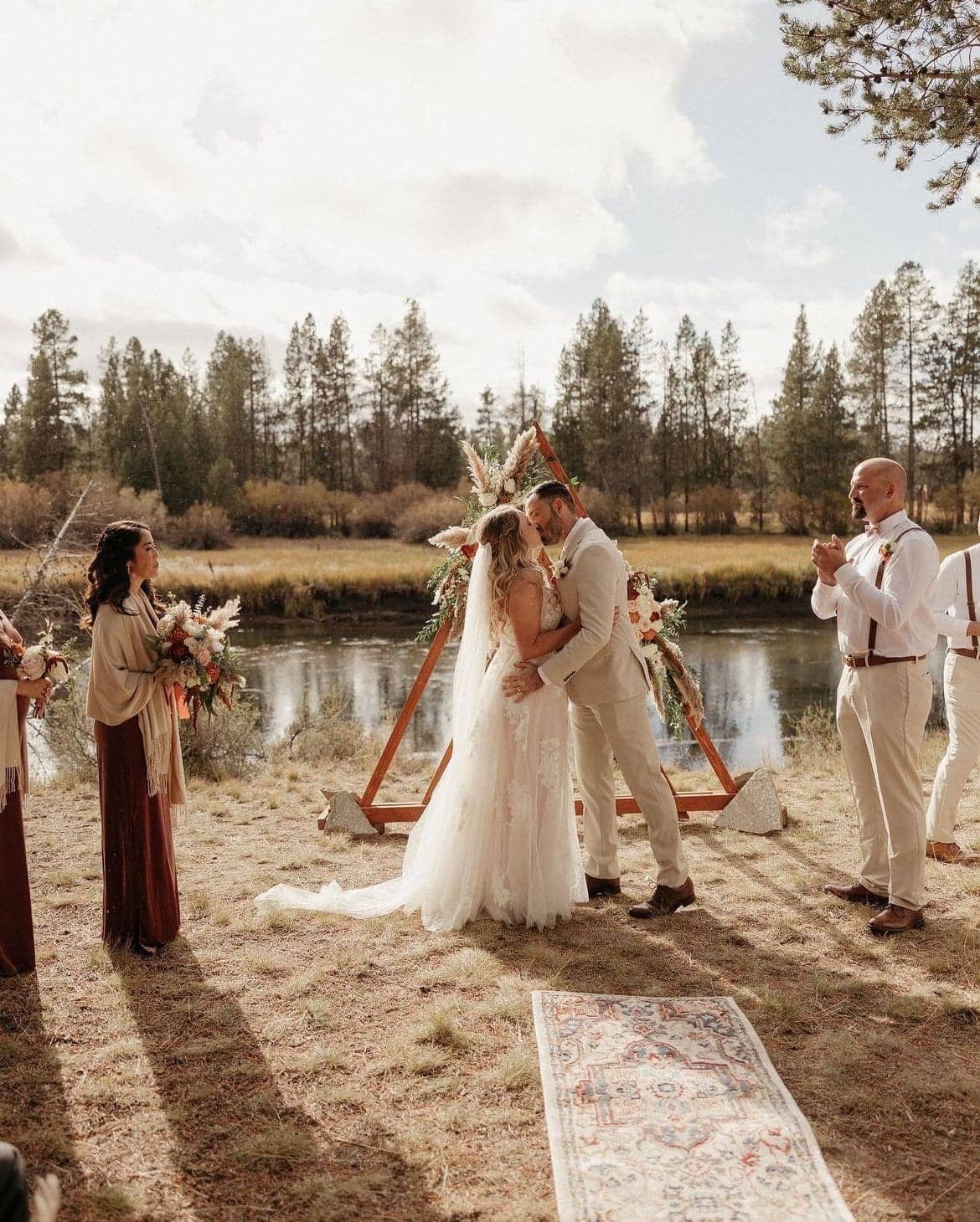 A couple kissing beneath a triangular floral arch at their riverside ceremony, attendants either side, Deschutes Riverfront Retreat near Sunriver, Oregon