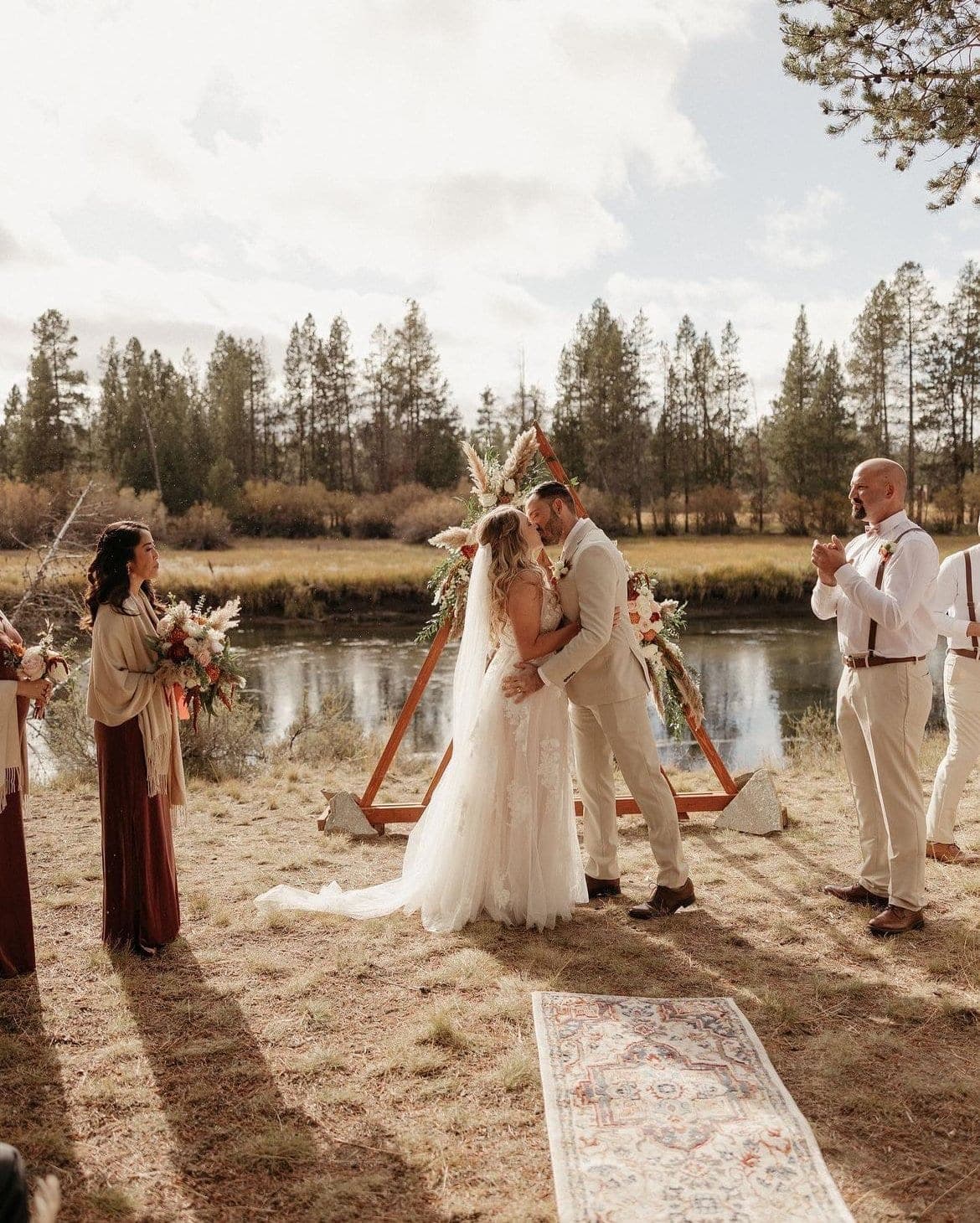 A couple kissing beneath a triangular floral arch at their riverside ceremony, attendants either side, Deschutes Riverfront Retreat near Sunriver, Oregon